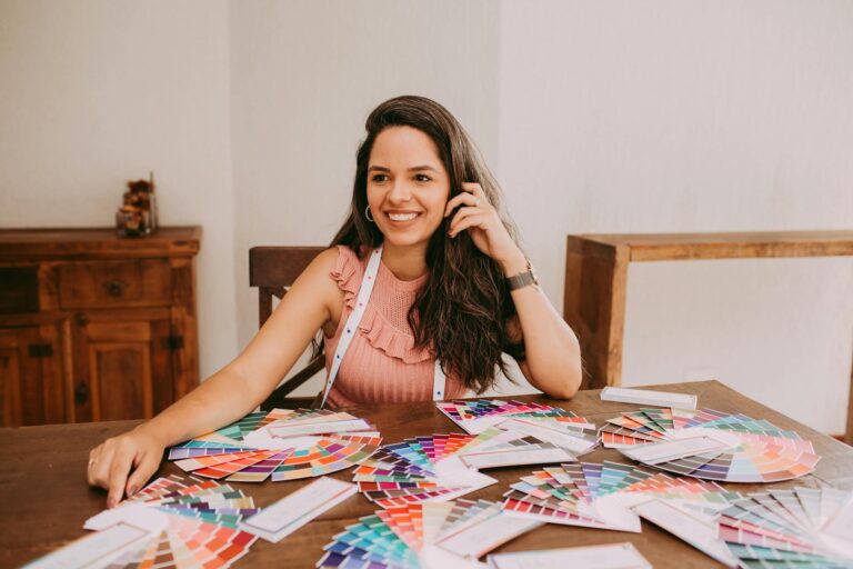 A woman with long hair, smiling while sitting at a table with various color swatches, suggesting creativity and design.