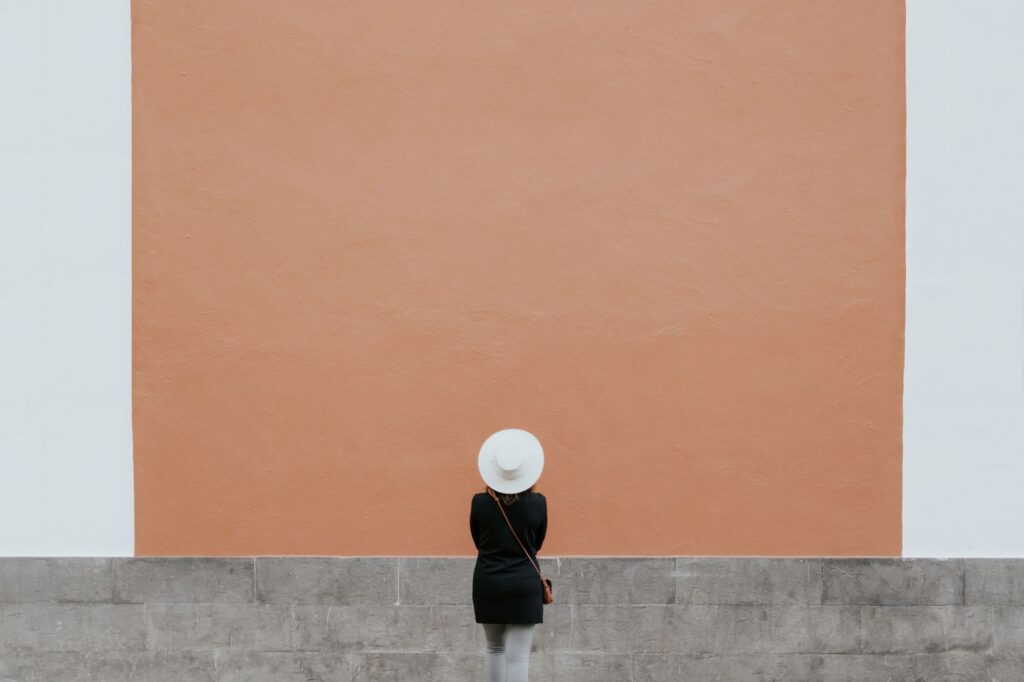 A minimalistic photo of a person in a hat against a large orange wall, perfect for design needs.