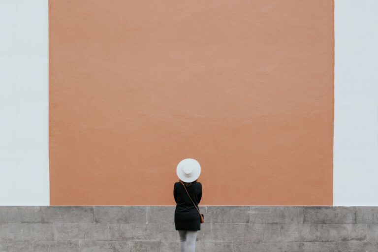 A minimalistic photo of a person in a hat against a large orange wall, perfect for design needs.