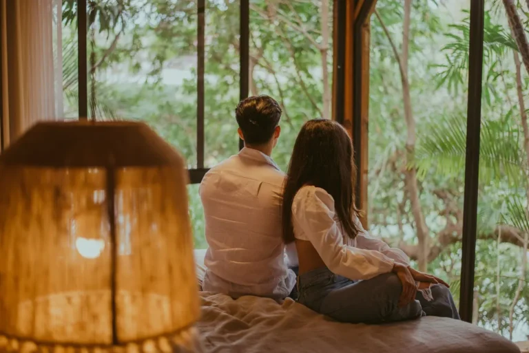 A couple sits on a bed in a resort bedroom, enjoying a tropical view.