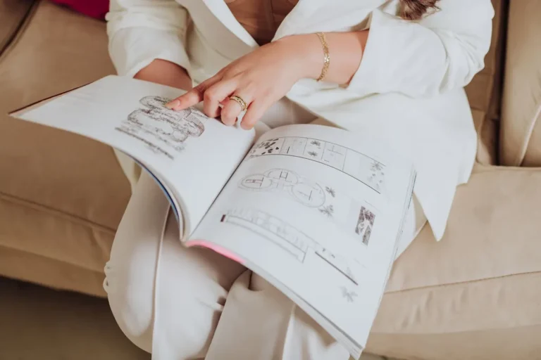 A woman in elegant attire reviewing architectural designs in a magazine while sitting on a sofa.