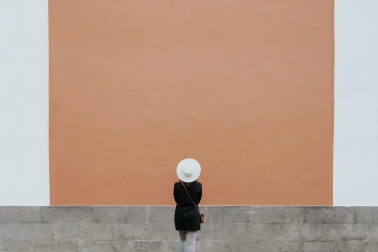 A minimalistic photo of a person in a hat against a large orange wall, perfect for design needs.