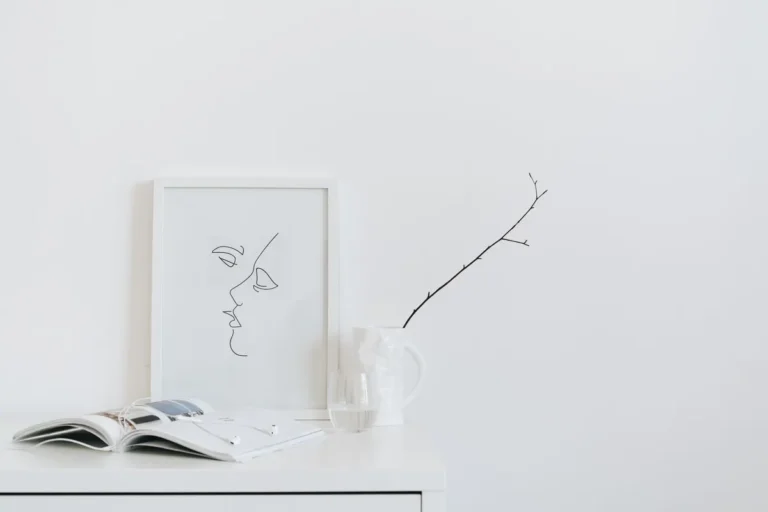 A serene minimalist desk setup featuring art, book, and glass on a white surface.